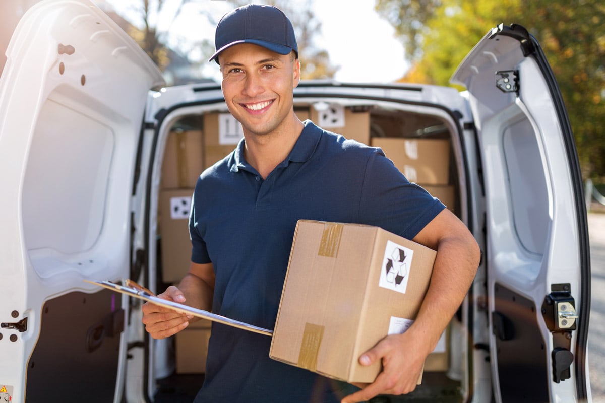 Smiling delivery driver holding a package and clipboard in front of an open van filled with boxes. 