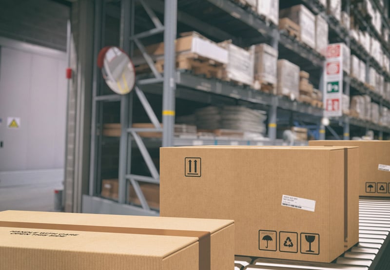 Cardboard boxes on a conveyor belt inside a warehouse with shelving and inventory in the background. 