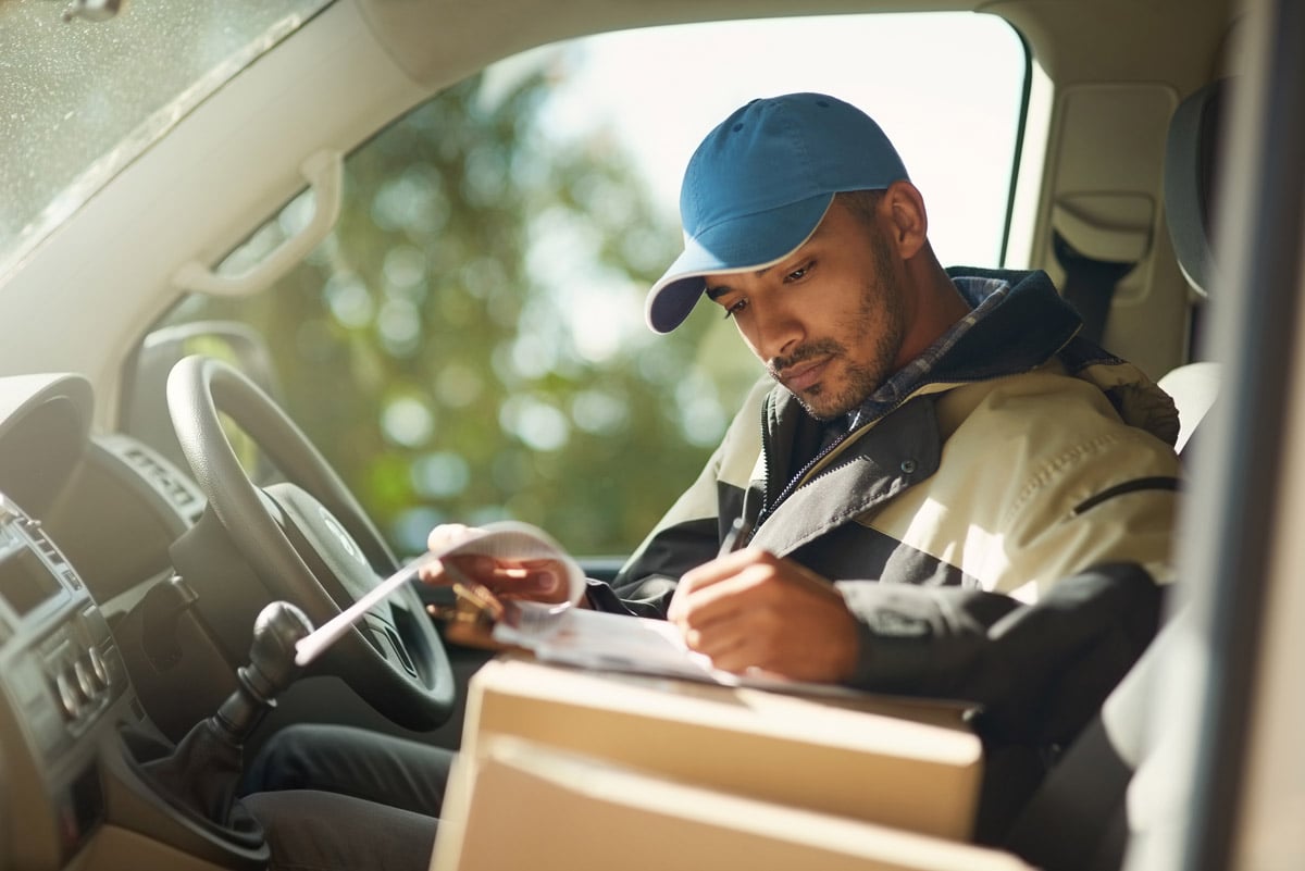 Delivery driver sitting in a van reviewing paperwork with packages on the seat beside him. 