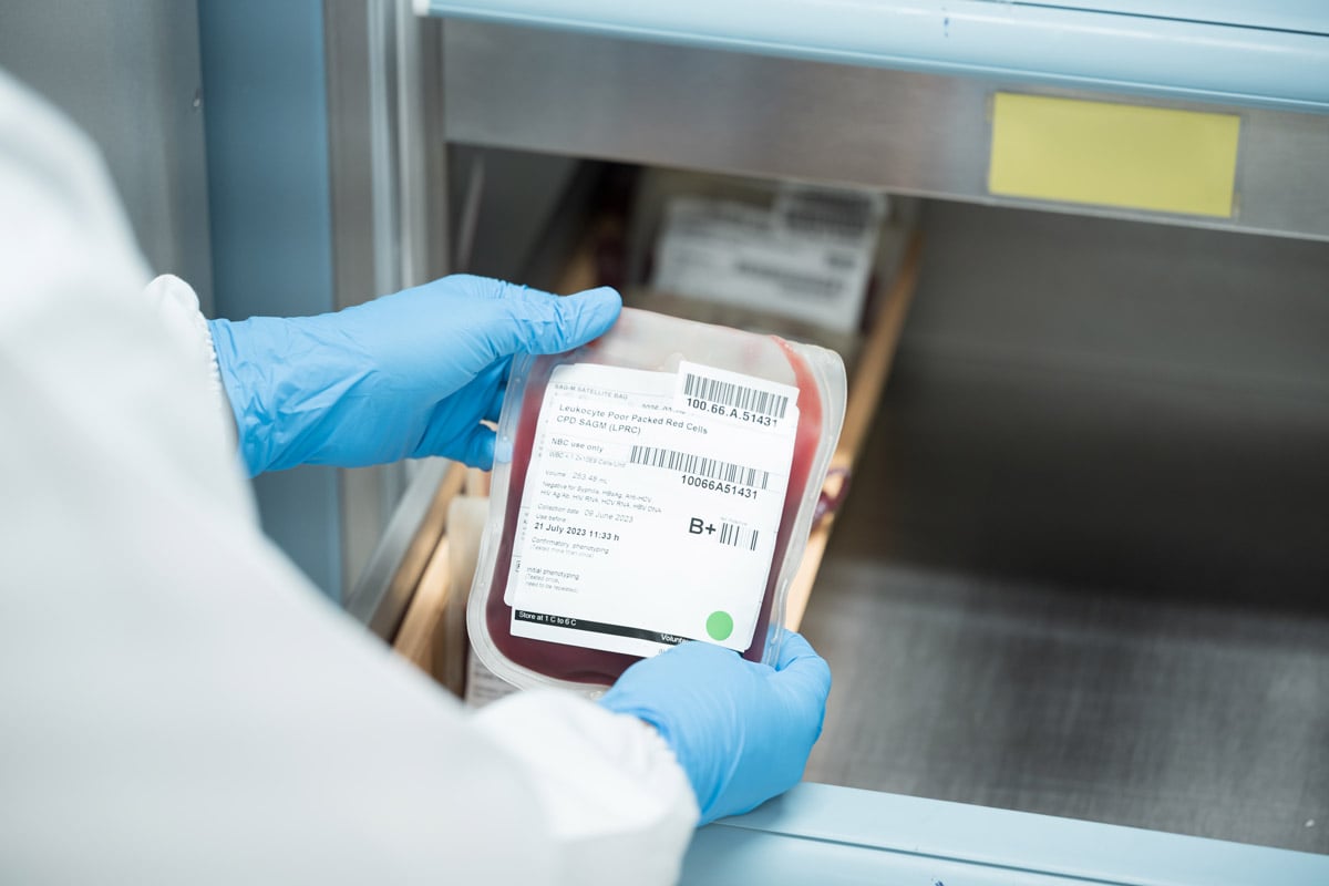 Gloved hands handling a labeled blood bag being placed into or removed from a medical storage unit. 