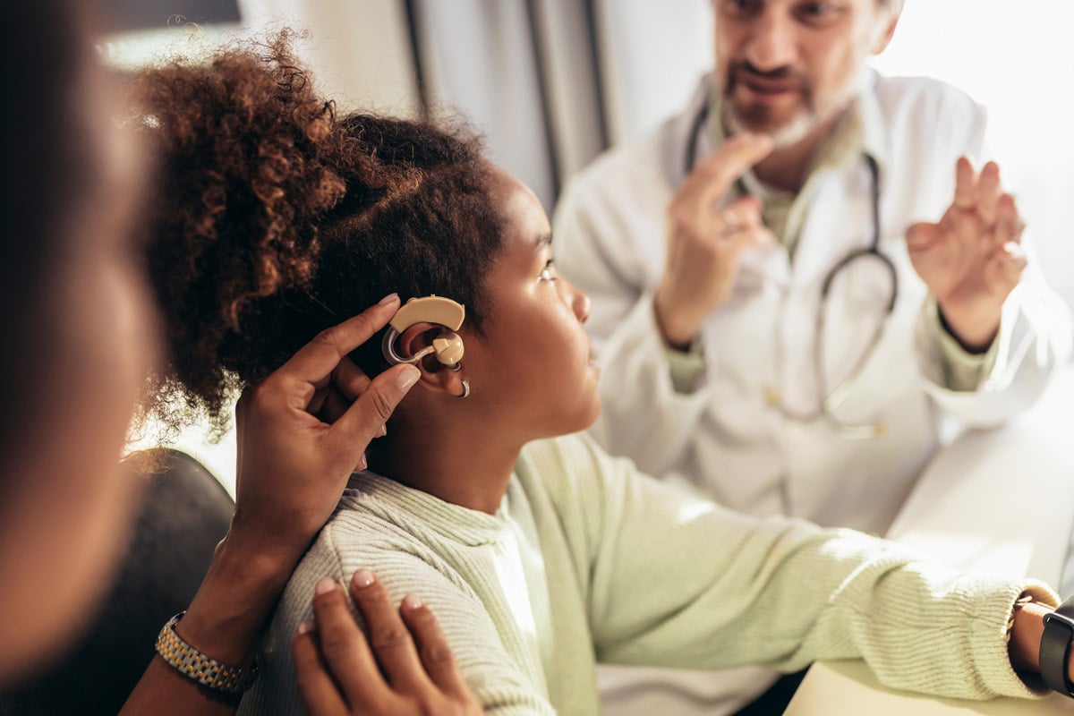 Child having a hearing aid fitted by a caregiver while a doctor provides guidance in a medical setting. 