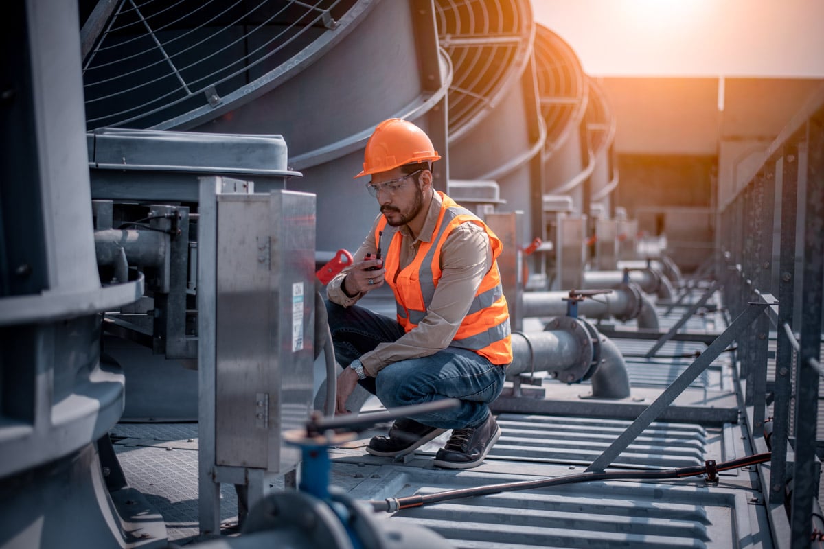 Industrial technician in safety gear inspecting and communicating via radio while checking rooftop mechanical equipment.
