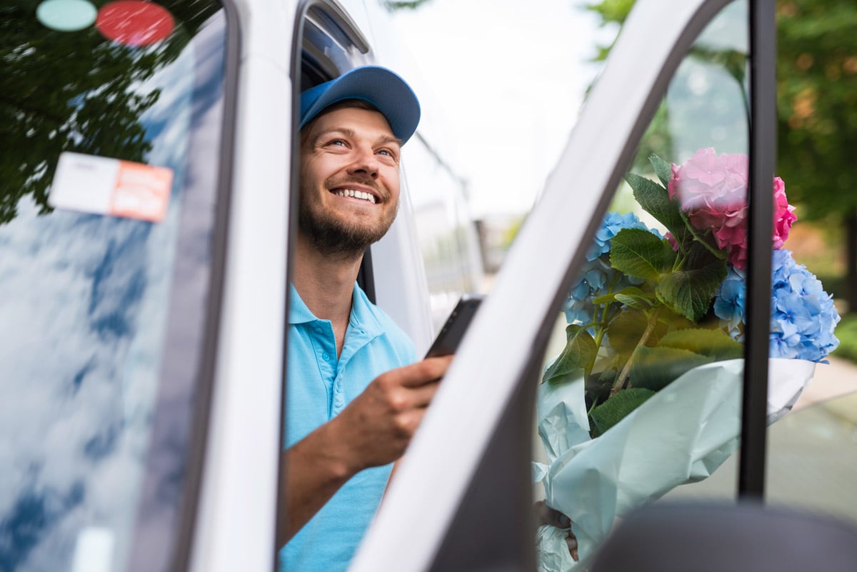 Smiling delivery driver holding a smartphone in a van with a bouquet of flowers, suggesting a floral delivery.